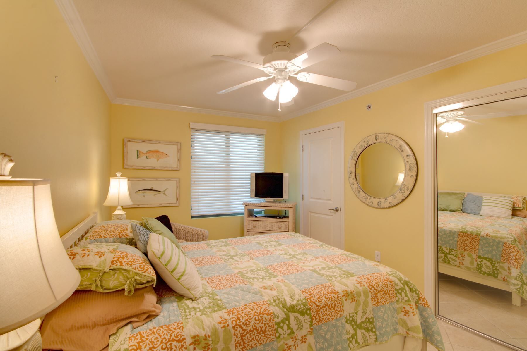 A bedroom with a bed, a television on a stand, a ceiling fan, a window with blinds, and a mirror on the wall.