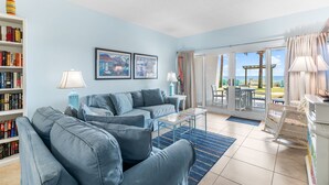 A living room with a sofa, coffee table, and a bookshelf. There is a large window with a view of the outdoors. - Sandy Key 128 Ground Floor Condo- Beachfront- NO HASSLE CHECK OUT (Pensacola)