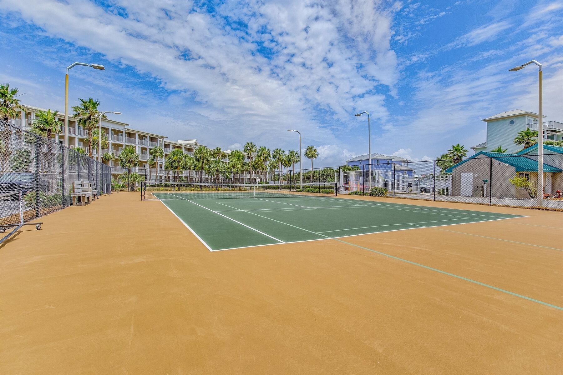 Ein Tennisplatz im Freien mit Netz, umgeben von einem Zaun, neben einem Gebäude mit Balkonen und Palmen.