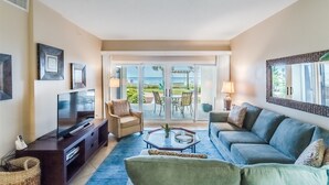A living room with a TV, sofa, and coffee table. There is a balcony with a view of the ocean. - Sandy Key 131 Ground Floor Condo- Beachfront- NO HASSLE CHECK OUT (Pensacola)