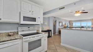 A modern kitchen with white cabinetry, a built-in microwave, and a stove. There is a dining area with a view of the ocean.
