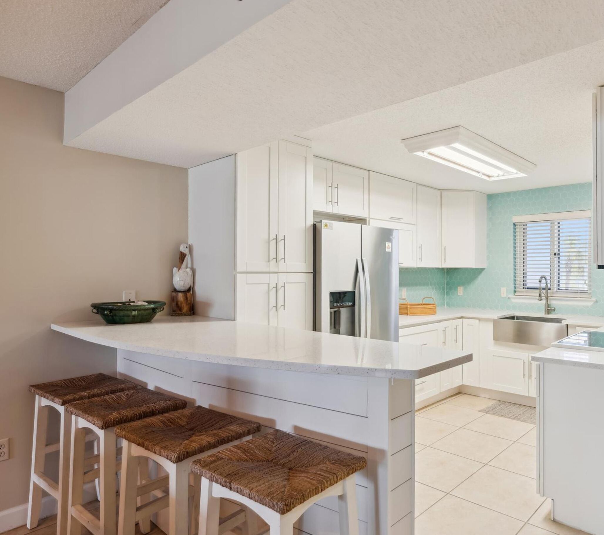A modern kitchen with white cabinetry, a central island with bar stools, a stainless steel refrigerator, and a window with blinds.