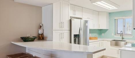 A modern kitchen with white cabinetry, a central island with bar stools, a stainless steel refrigerator, and a window with blinds.
