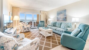 A spacious living room with a large window, a sofa set, a television, and a dining area with a table and chairs.