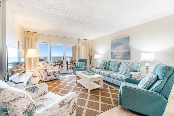 A spacious living room with a large window, a sofa set, a television, and a dining area with a table and chairs. - Sandy Key 233 Second floor -Beachfront view -NO HASSLE CHECK OUT (Perdido Key)