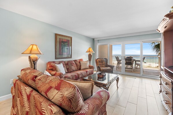A spacious living room with a sofa set, a coffee table, and two lamps. There is a large window with a view of the beach.