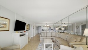 Un salon moderne avec un canapé d’angle, un téléviseur à écran plat et une cuisine en arrière-plan.