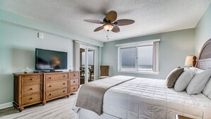 Une chambre à coucher avec un lit, un téléviseur posé sur une commode en bois, un ventilateur de plafond et une fenêtre donnant sur l’océan.