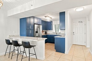 A modern kitchen with blue cabinets, a stainless steel refrigerator, a built-in oven, and a bar area with stools.