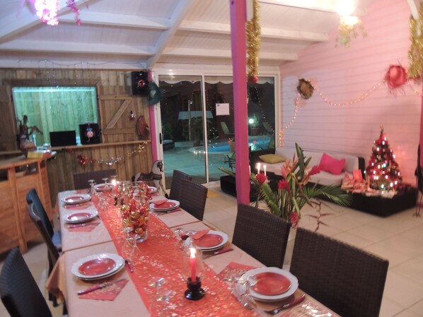 A dining area with a long table set for a meal, a Christmas tree, and a pool in the background.