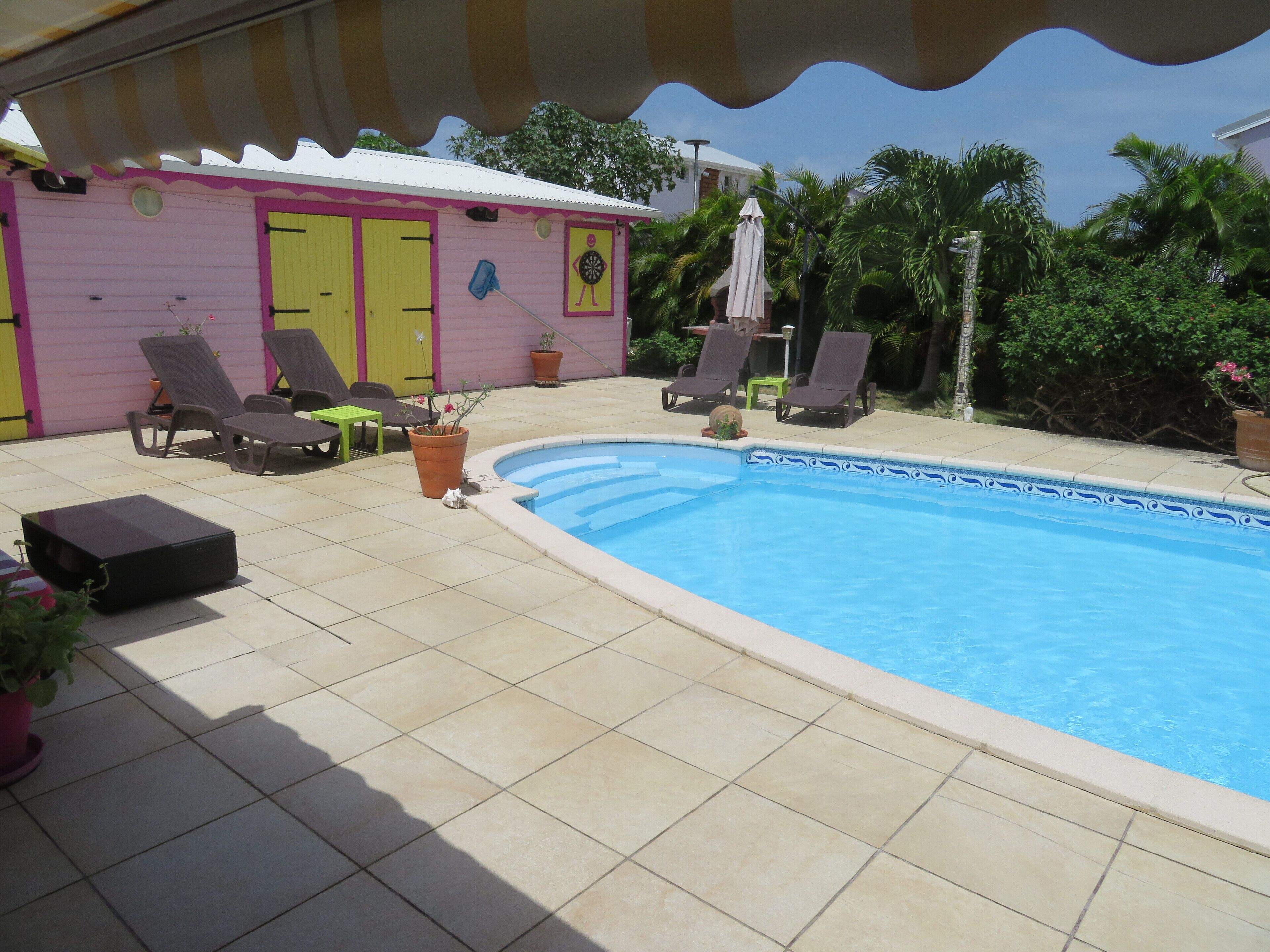 Un espace piscine doté d’une terrasse carrelée, de chaises longues, d’une petite table et d’un bâtiment aux portes jaunes.