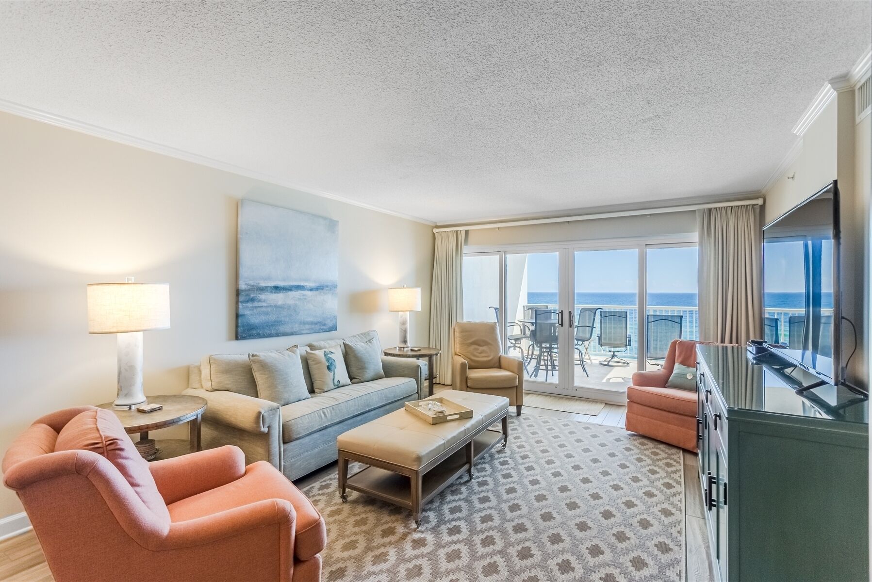 A hotel room with a sofa, armchairs, a coffee table, a TV, and a balcony with a view of the ocean.