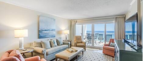 A hotel room with a sofa, armchairs, a coffee table, a TV, and a balcony with a view of the ocean.