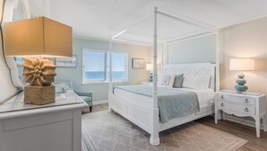 A four-poster bed with a white headboard, a bedside table with a lamp, and a chair with a cushion. - Sandy Key 516 5th floor -Beachfront view -NO HASSLE CHECK OUT (Pensacola)