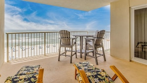 A balcony with two chairs and a table, overlooking a beach with umbrellas and people.