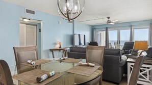 A dining area with a glass table, woven placemats, and a chandelier. There are two sofas and a television in the background. - Sandy Key 621 6th floor -Beachfront view -NO HASSLE CHECK OUT (Pensacola)
