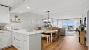 A modern kitchen with white cabinetry, a dining area with a wooden table and wicker chairs, and a view to the outside.