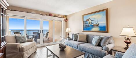 A living room with a sofa, armchair, and coffee table. There is a painting on the wall and a view of the ocean through the glass doors.