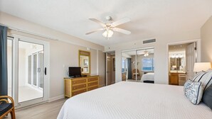 A hotel room with a large bed, a ceiling fan, a TV on a wooden dresser, a bathroom with a mirror and sink, and a sliding glass door leading to a balcony. - Sandy Key 735 -7th floor -Beachfront view -NO HASSLE CHECK OUT (Pensacola)