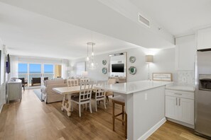 A modern kitchen with white cabinetry, a central island, and a dining area with a white table and chairs. - Sandy Key 827 -8th floor -Beachfront view -NO HASSLE CHECK OUT (Pensacola)