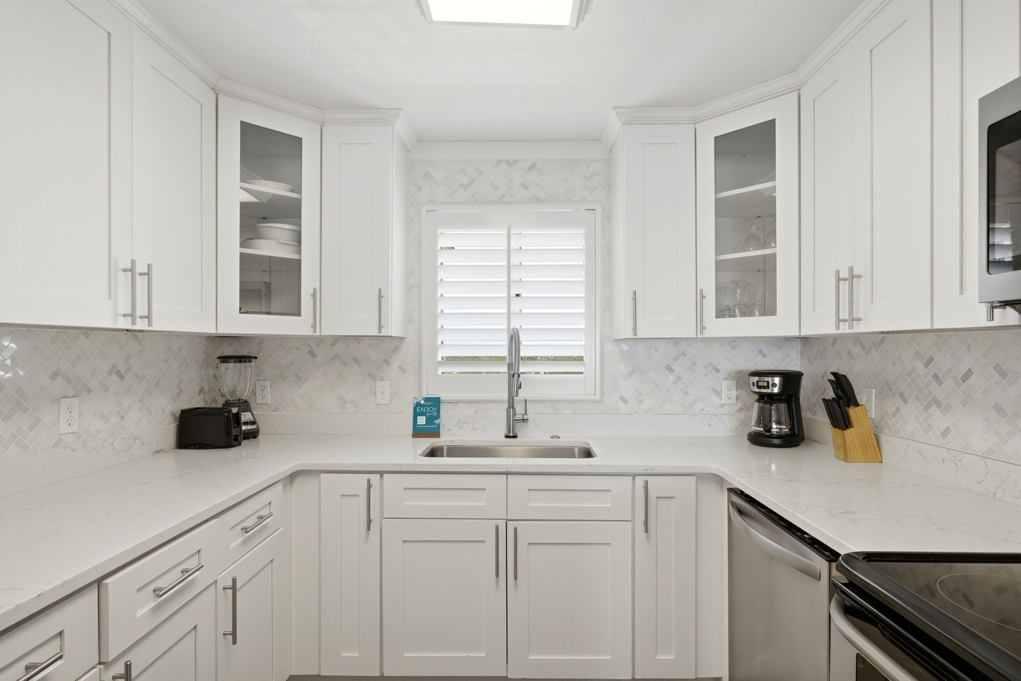 A modern kitchen with white cabinetry, a built-in stove, a sink, and a window with blinds.