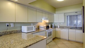 Una cocina con armarios blancos, encimera de granito, microondas, estufa, lavavajillas y una ventana con persianas.