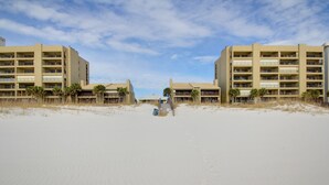 Un hotel frente a la playa con varios pisos, una playa arenosa en primer plano y palmeras.