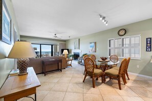 A spacious living room with a sofa, a dining table with chairs, a fireplace, and a view of the ocean through large windows.