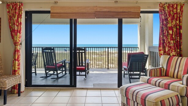 A room with a sliding glass door leading to a balcony with chairs and a view of the beach.