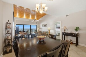 Un espace repas comprenant une grande table en bois, des chaises, un lustre et une vue dégagée sur l’extérieur grâce à une grande fenêtre.