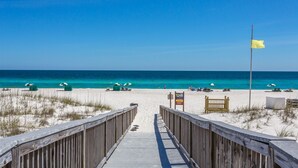 A wooden boardwalk leading to a beach with clear blue water, a yellow flag, and beach umbrellas. - Spanish Key 207  Beachfront view  - NO HASSLE CHECK OUT (Perdido Key)