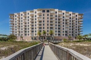 Un hotel de varios pisos con balcones, un paseo marítimo que conduce a la entrada y palmeras en primer plano.