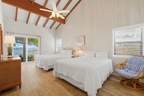 Una habitación con dos camas, una silla de mimbre, un ventilador de techo y vista al mar a través de un ventanal corredero.