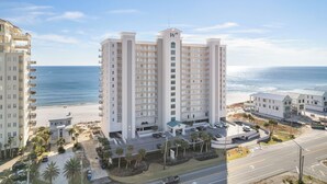 A high-rise hotel with multiple balconies, a parking area, and a view of the beach.