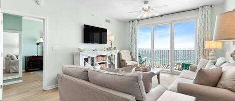 A spacious living room with a large sofa, a coffee table, and a TV. There is a balcony with a view of the city.