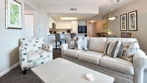 Een woonkamer met een bank, fauteuil en salontafel. Achtergrond: een keuken met witte kastjes en een eethoek met stoelen.