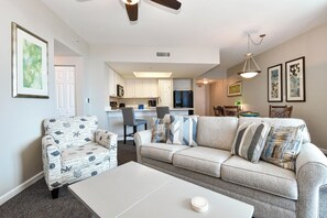 Ein Wohnzimmer mit einer Couch, einem Sessel und einem Couchtisch. Im Hintergrund befindet sich eine Küche mit weißen Schränken und ein Essbereich mit Stühlen.