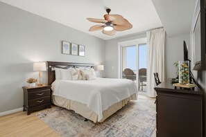 A bedroom with a bed, bedside tables, a ceiling fan, a large window with curtains, and a view of the outdoors.