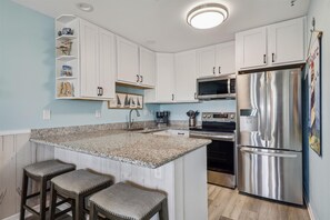 Eine Küche mit weißen Schränken, einer Granit-Arbeitsplatte, einem Edelstahl-Kühlschrank und einer Mikrowelle. Zwei Barhocker mit gepolsterten Sitzen sind vorhanden.
