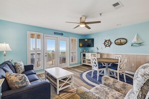 Ein Wohnzimmer mit Fernseher, Deckenventilator und Blick auf den Strand durch die Glastüren.