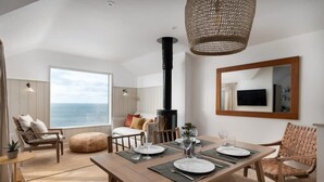 Un coin repas avec une table en bois dressée pour quatre personnes, une grande fenêtre donnant sur la mer et une cheminée noire.