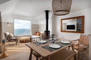 Uma sala de jantar com uma mesa posta para quatro pessoas, uma janela grande com vista para o mar, uma lareira e uma televisão fixada na parede.