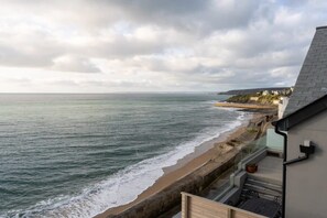 A coastal view from a balcony overlooking a beach with waves, a stone wall, and a building with a sloped roof.
