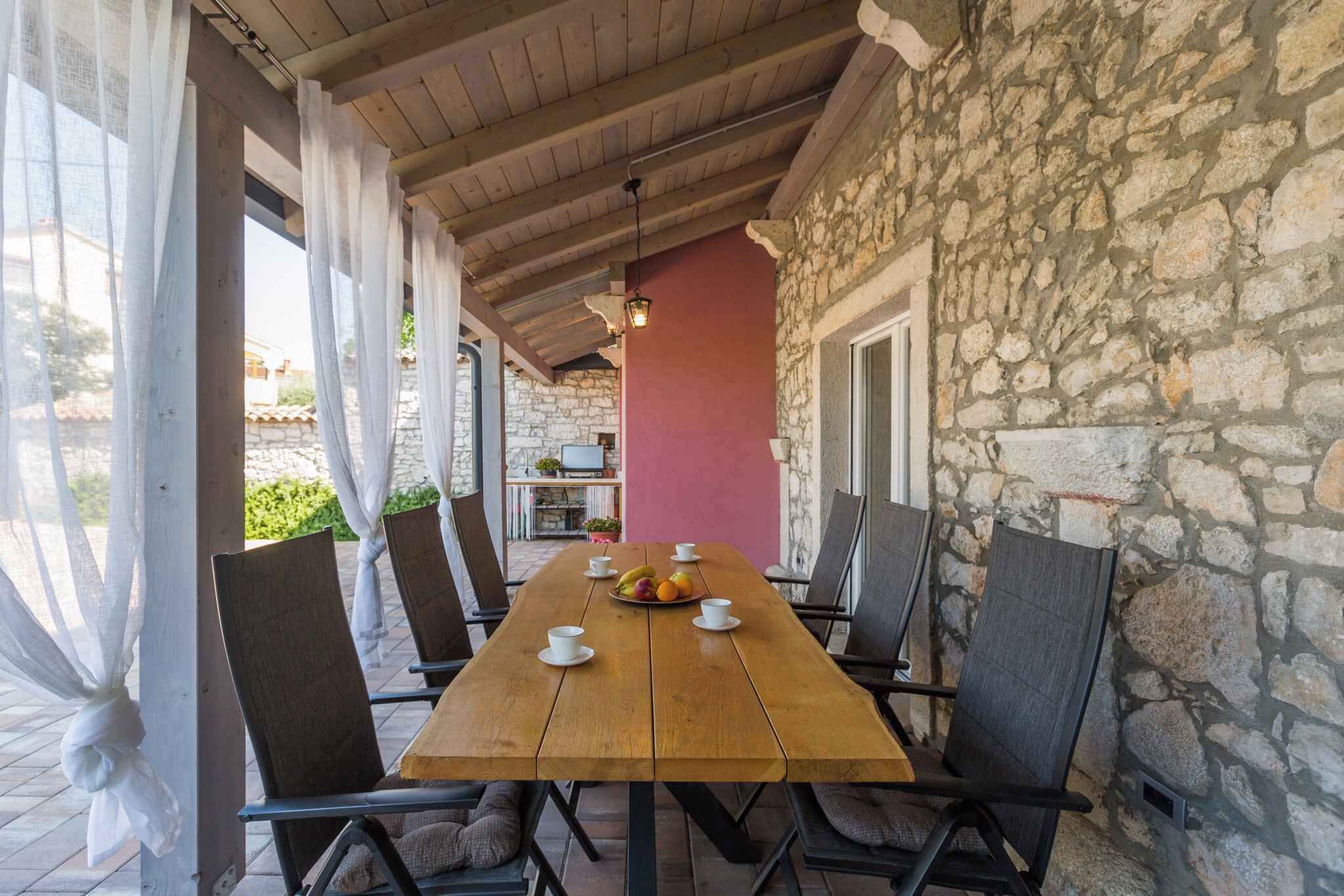 A wooden table with chairs, a fruit bowl, and cups, set in a covered outdoor area with stone walls and a red wall in the background.