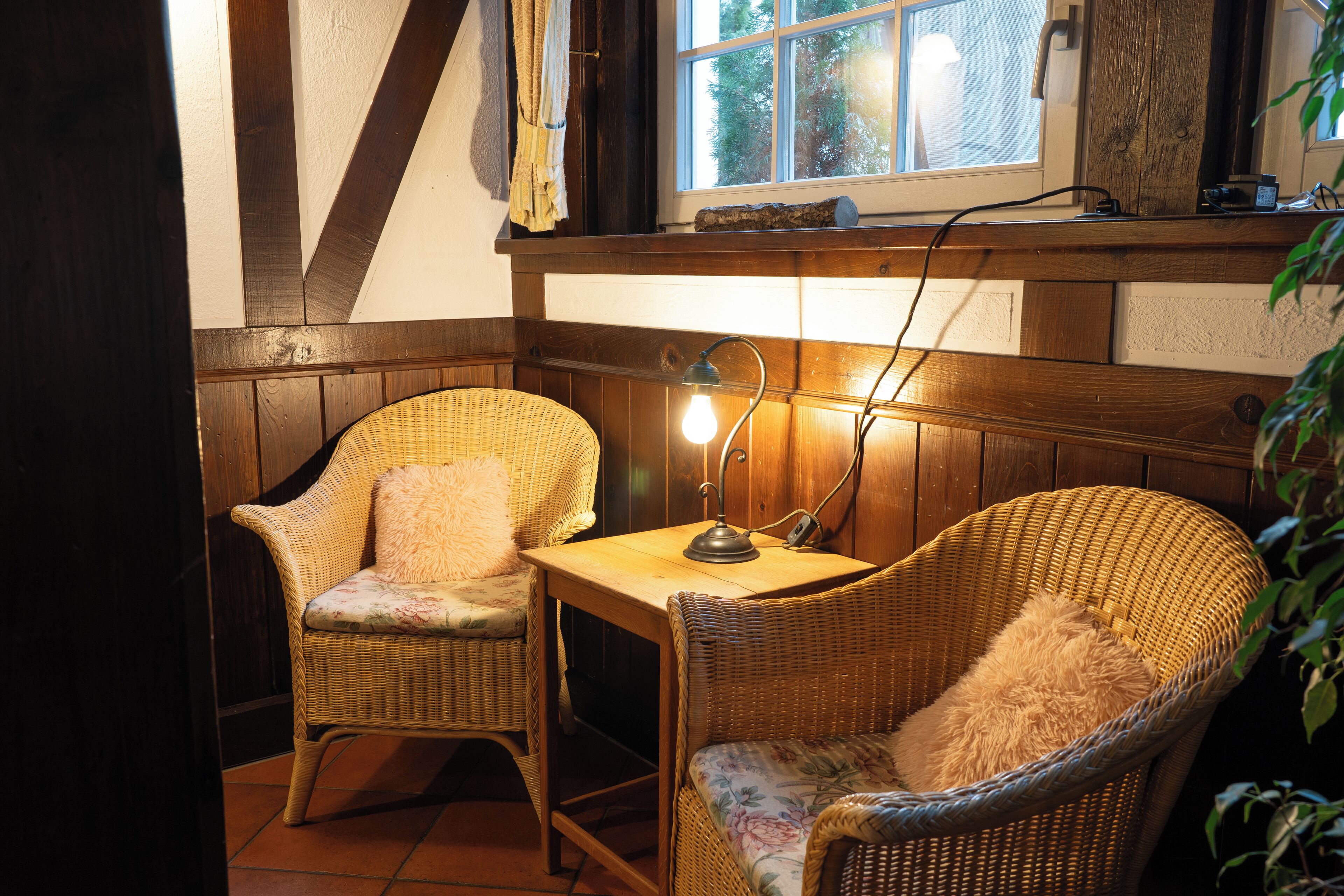 Une pièce d’angle avec deux fauteuils en osier, une petite table avec une lampe et une fenêtre donnant sur des arbres.