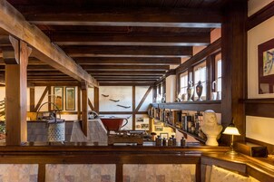 Um interior rústico com vigas de madeira aparente, balcão de madeira e vários objetos decorativos, incluindo um busto e quadros nas paredes.