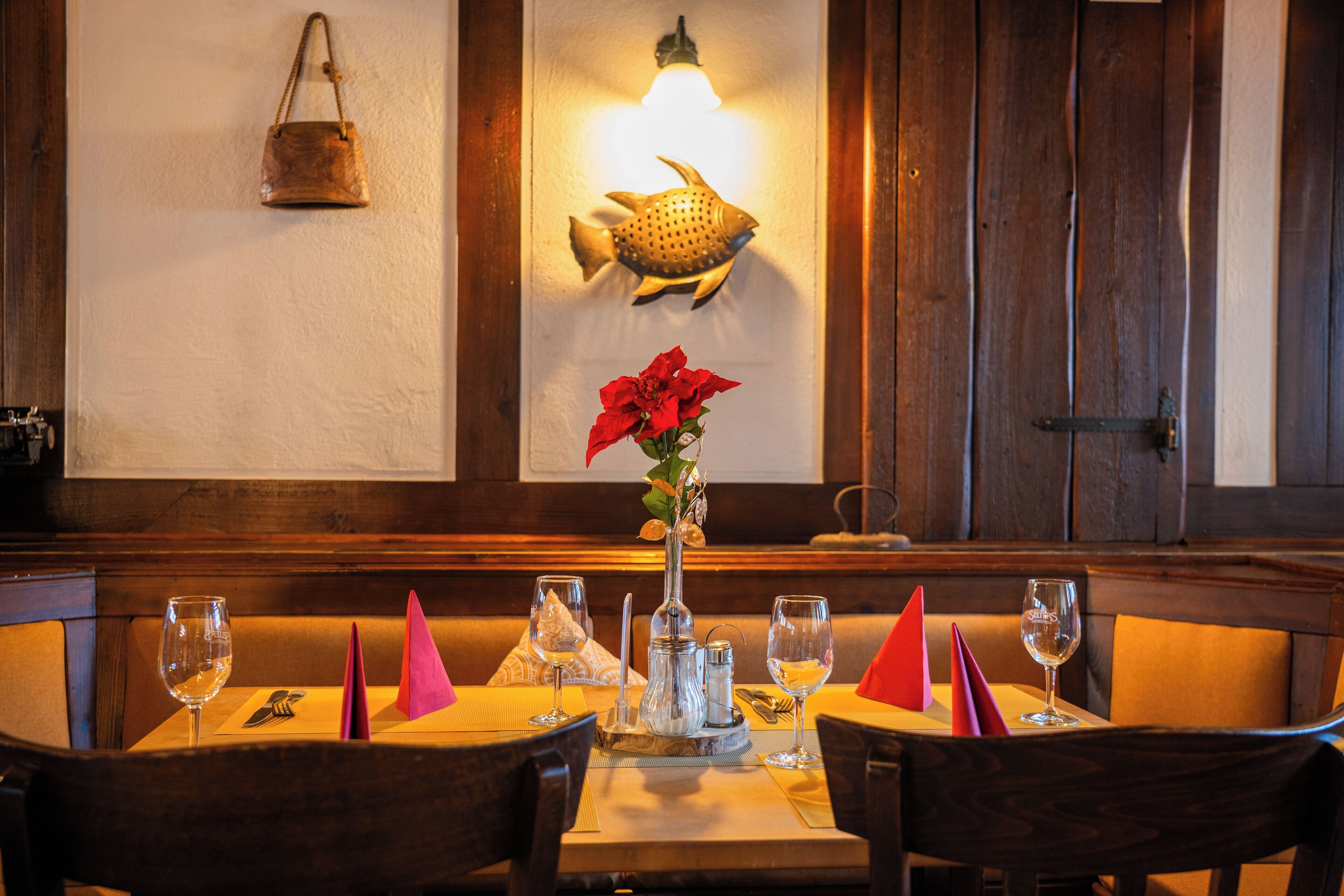 A restaurant interior with wooden paneling, a fish-shaped wall light, a vase with red flowers, and neatly arranged tables with wine glasses and menus.