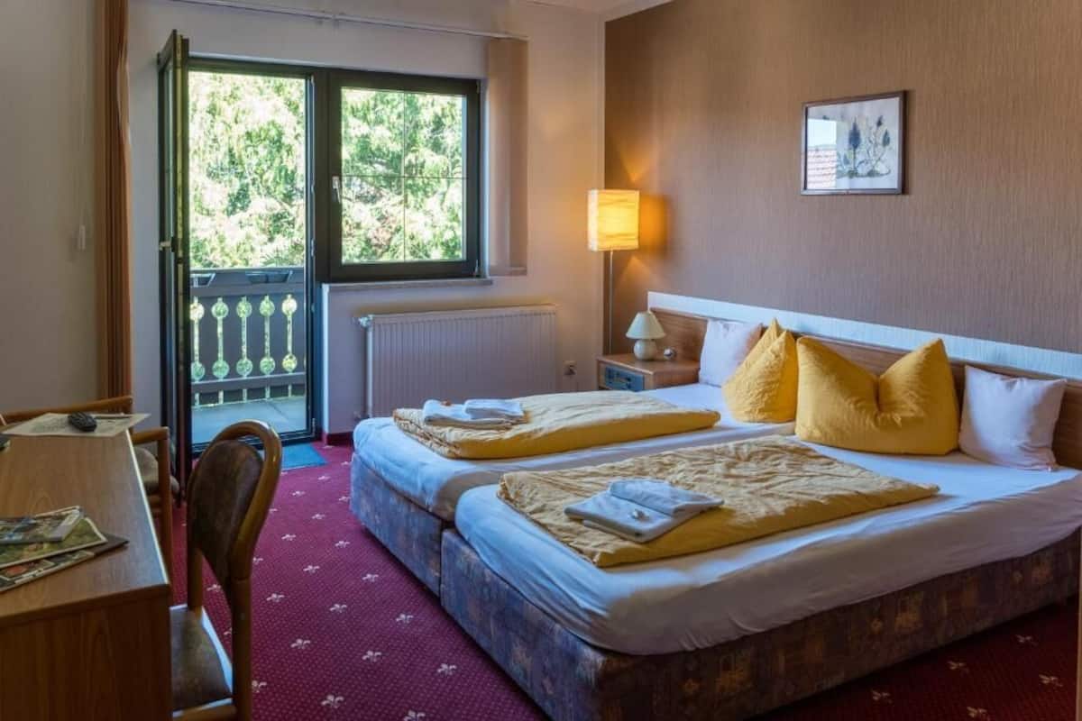 Habitación de hotel con dos camas, un escritorio, una silla, una ventana con vistas a árboles y una lámpara.