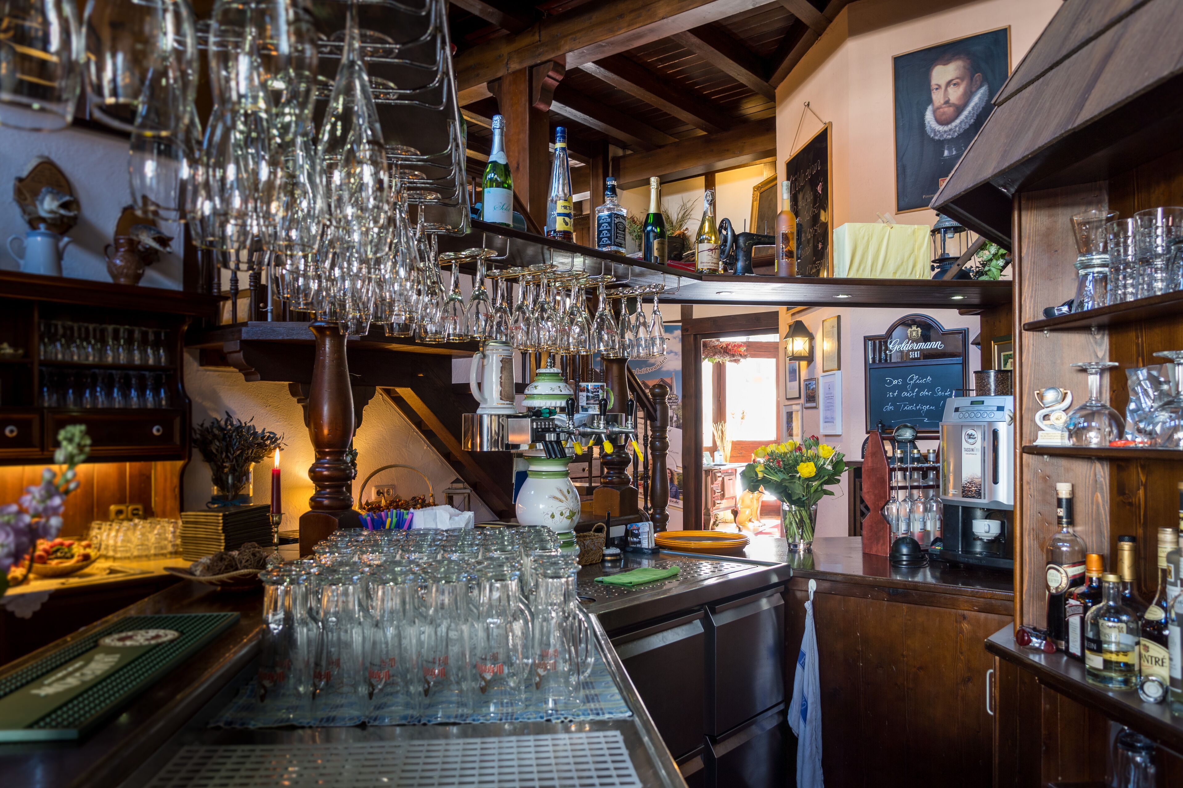 Zona de bar con numerosos vasos colgando del techo, una variedad de botellas y copas en estantes, y una máquina de café.