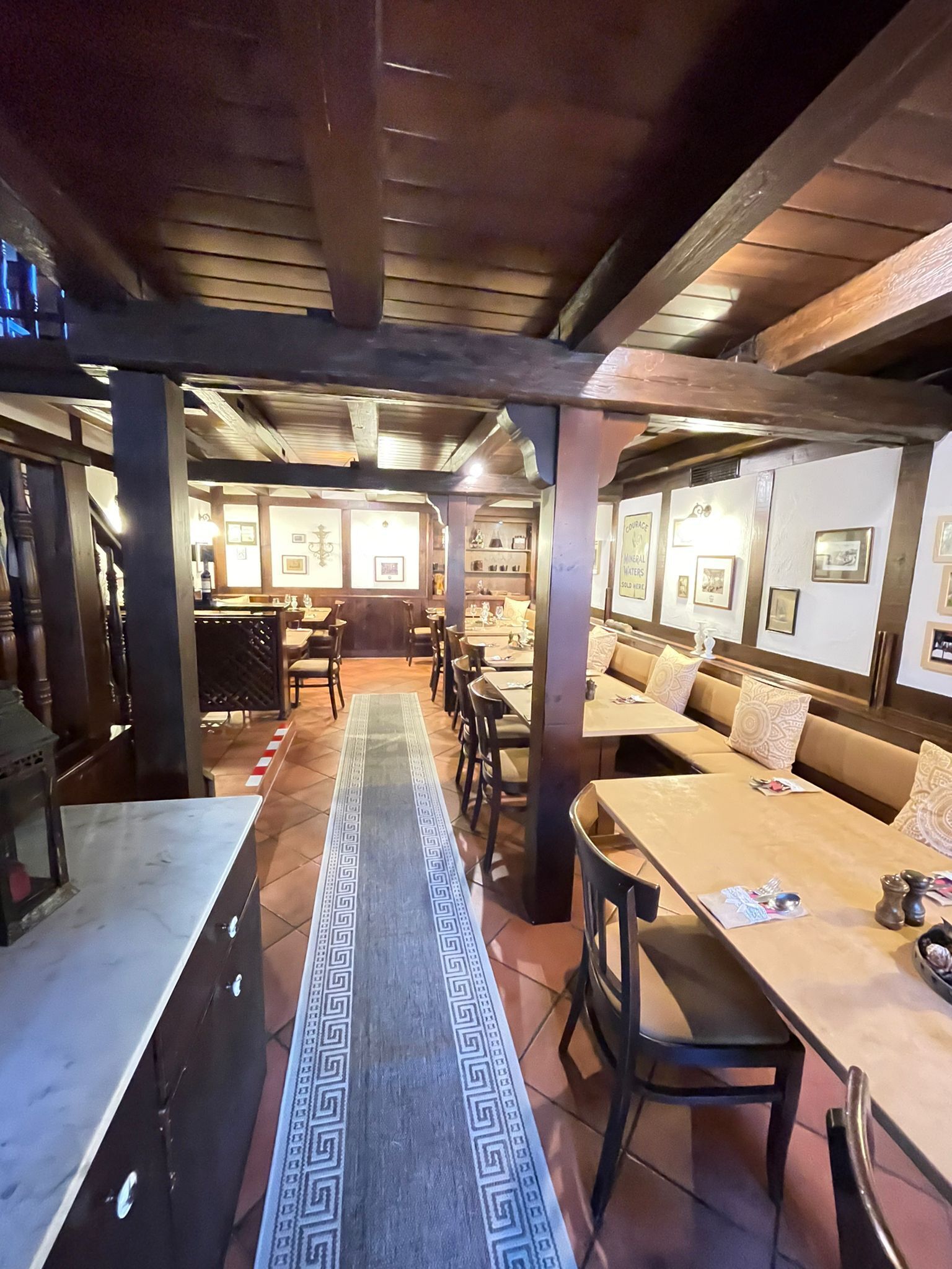 A restaurant with wooden beams, a long table with a patterned runner, and framed pictures on the walls.